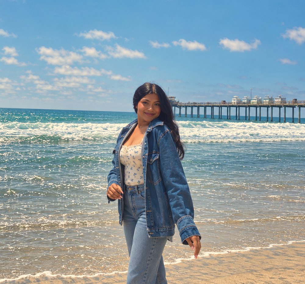 Asha Krishna, founder of Aloha Girl Cosmetics, smiling on the beach by the pier in a denim jacket